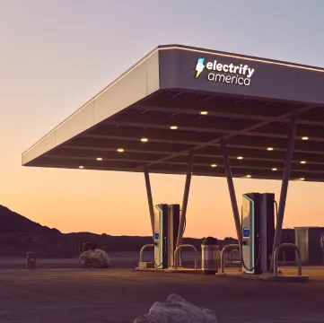 An Electrify America charging station lit up at sunset, two chargers are visible under a large open-air shelter. In the background is a large rock and the start of a mountain in the distance.