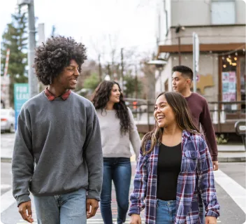 Four people walking in pairs of two towards the camera crossing a street, each pair is looking towards each other and smiling.