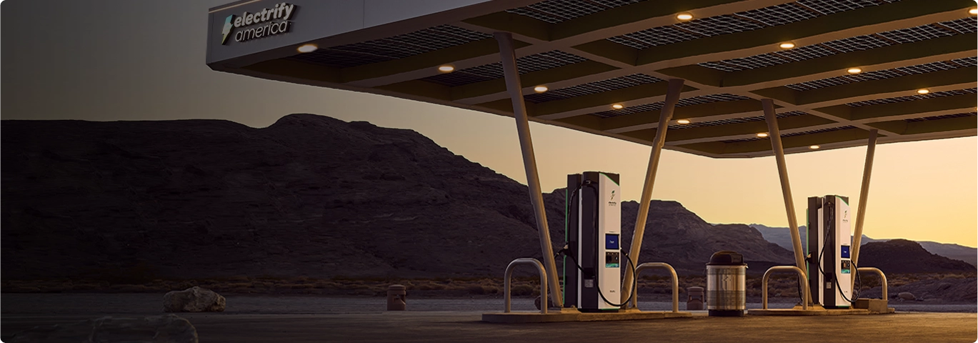 An Electrify America charging station lit up at dusk, four chargers are lined up in two rows of two under a large open-air shelter. In the background is the side of a large mountain and purple darkening sky.