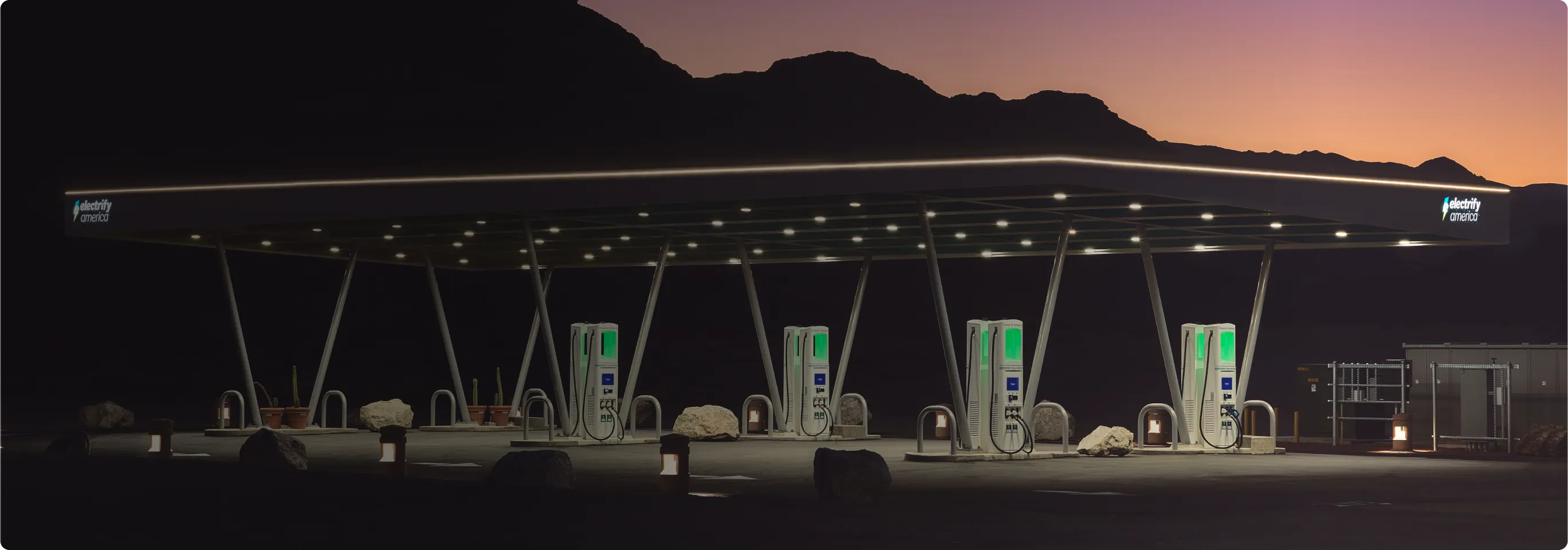 An Electrify America charging station lit up at dusk, four chargers are lined up in two rows of two under a large open-air shelter. In the background is the side of a large mountain and purple darkening sky.