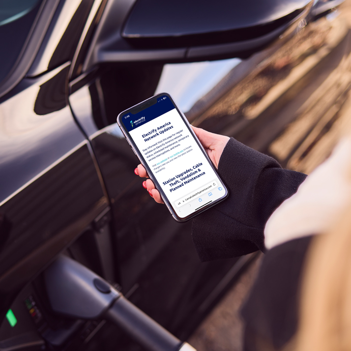 Person holding a cell phone open to the electrify america site next to a vehicle charging.