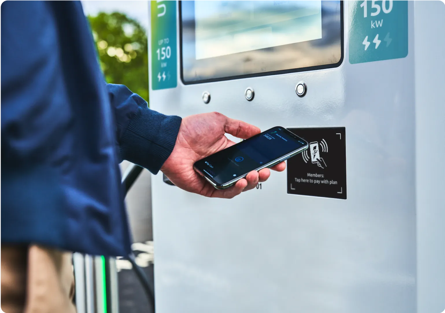 This image displays a person holding their phone up to an NFC reader on an Electrify America charger.